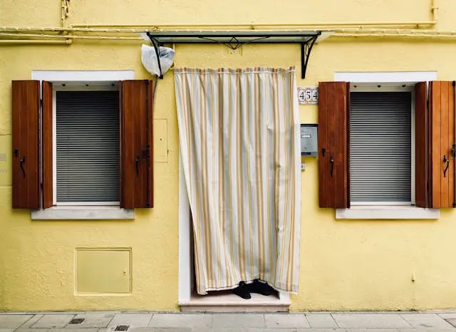 A yellow house exterior with wooden shutters and striped curtains at the entrance.