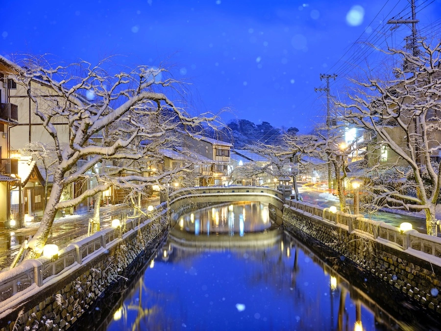 Kinosaki Onsen town dusted in winter snow, with traditional wooden buildings lining a quiet canal.