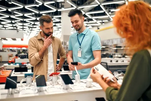A store employee helps two customers look at smartphones displayed on a counter.