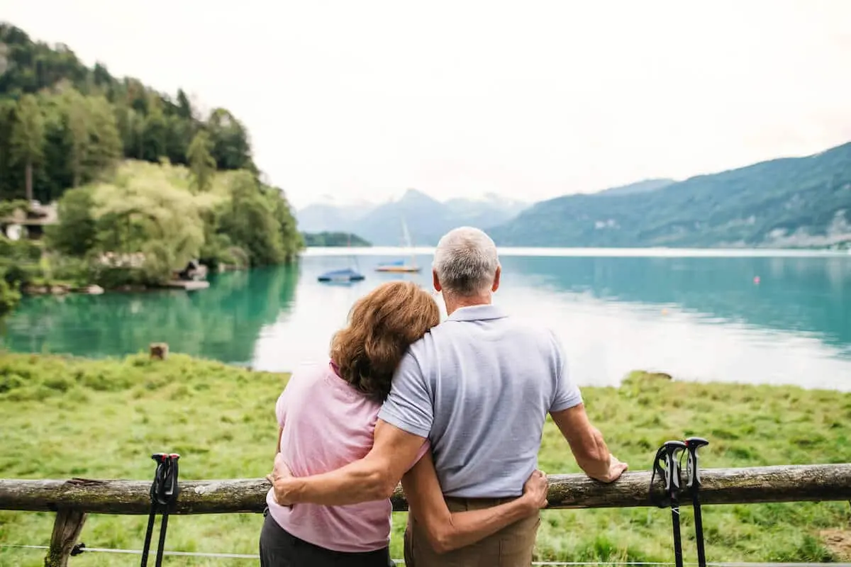 An older couple embraces while looking at a calm lake surrounded by mountains and trees.